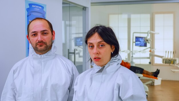 Retrato del médico del equipo en el consultorio dental quitándose las máscaras protectoras mirando a la cámara con marcas en la cara con traje de ppe en la sala de espera. Concepto de nueva clínica normal en brote de coronavirus