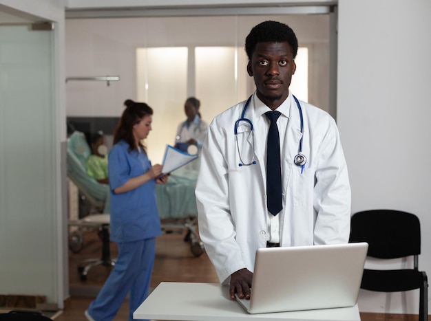 Retrato de médico especialista afroamericano de pie en el escritorio en la sala de hospital escribiendo experiencia en enfermedad en la computadora portátil. Hombre médico que trabaja en el tratamiento sanitario. Servicios de medicina