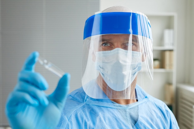 Retrato de médico irreconocible con máscaras protectoras y guantes sosteniendo ampolla con vacuna Covid-19