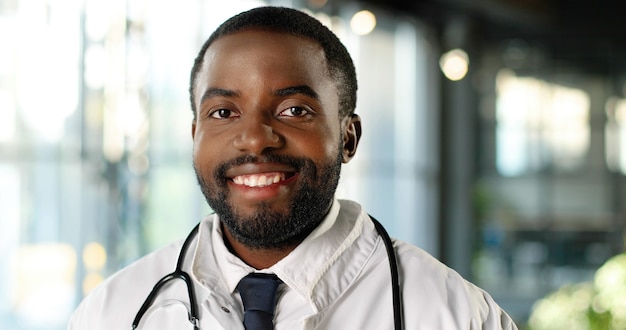 Retrato de médico joven afroamericano con estetoscopio sonriendo a la cámara con alegría. Sonrisa feliz guapo médico masculino. Médico en bata blanca en la clínica. Interior.