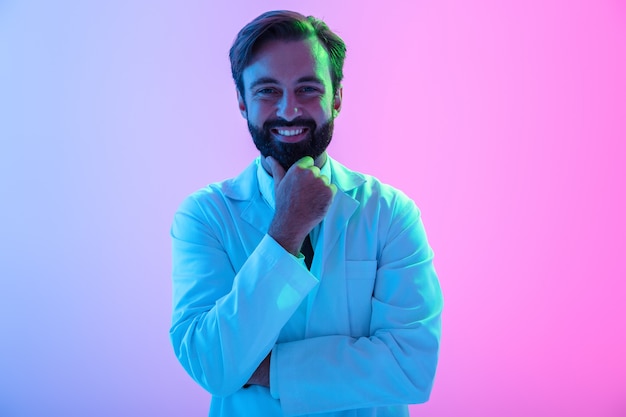 Retrato de un médico joven confiado en uniforme que se encuentran aisladas sobre neblina rosa-azul, mirando a la cámara