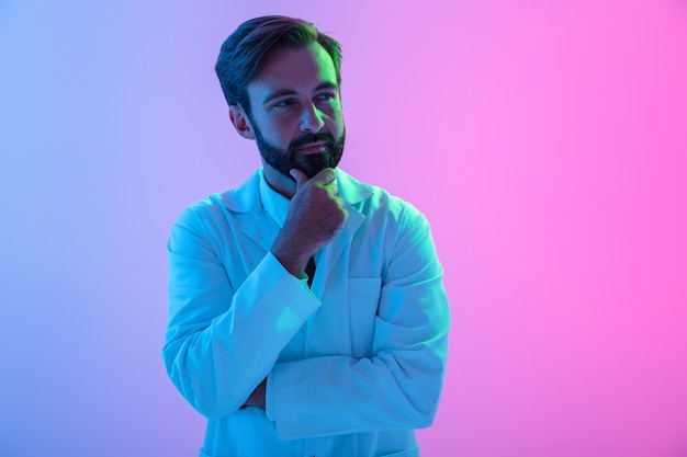 Retrato de un médico joven confiado en uniforme que se encuentran aisladas sobre neblina rosa-azul, mirando a otro lado