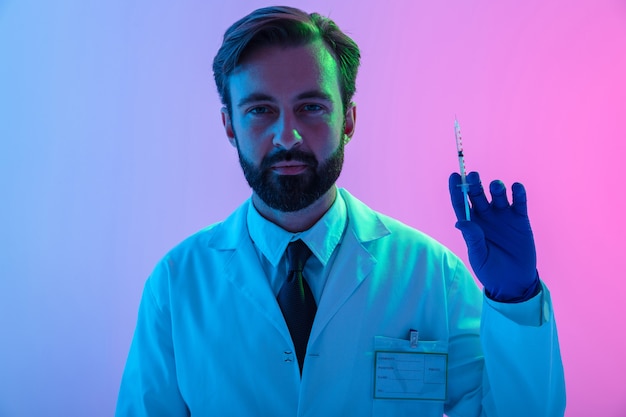 Retrato de un médico joven confiado en uniforme que se encuentran aisladas sobre neblina rosa-azul, sosteniendo una jeringa con líquido