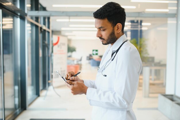 Retrato de un médico latino indio feliz y amistoso que lleva bata blanca con estetoscopio alrededor del cuello de pie en una clínica privada moderna mirando a la cámara Concepto de atención médica