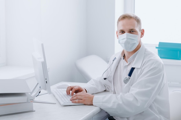 Retrato de un médico con una máscara protectora que está sentado en un escritorio de computadora en su oficina en la clínica Un cardiólogo exitoso en el trabajo