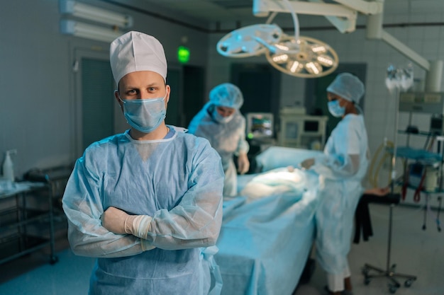 Retrato de un médico masculino con uniformes quirúrgicos y máscaras de pie posando mirando a la cámara con la mano doblada