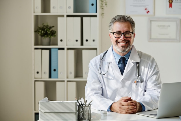 Retrato de un médico que tiene una cita en su oficina, se sienta a la mesa con una computadora portátil y sonríe a c