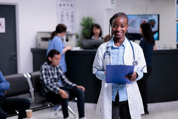 Retrato de un médico sonriente con estetoscopio en la concurrida recepción del hospital esperando la próxima cita con el paciente. Médico afroamericano posando confiado en bata de laboratorio y sosteniendo portapapeles.