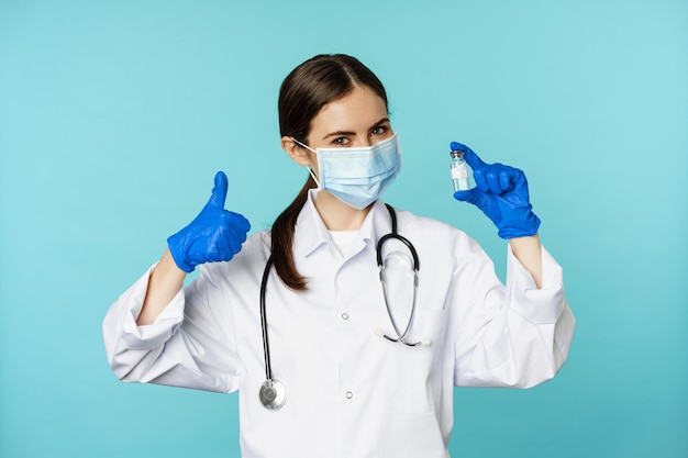 Retrato de un médico sonriente, personal médico con máscara médica facial y guantes de goma, mostrando los pulgares hacia arriba y el coronavirus, la vacuna omicron, de pie sobre fondo azul.