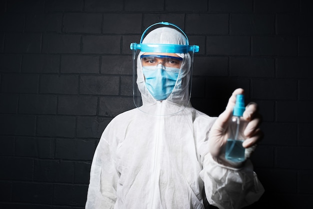 Retrato de médico con traje de PPE contra coronavirus y covid-19, sosteniendo una botella de desinfectante en aerosol contra una pared de ladrillo negro