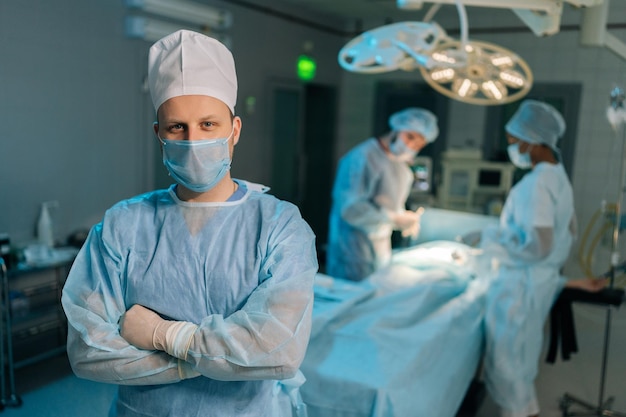 Retrato de un médico en uniforme quirúrgico y máscaras de pie posando mirando a la cámara con la mano doblada