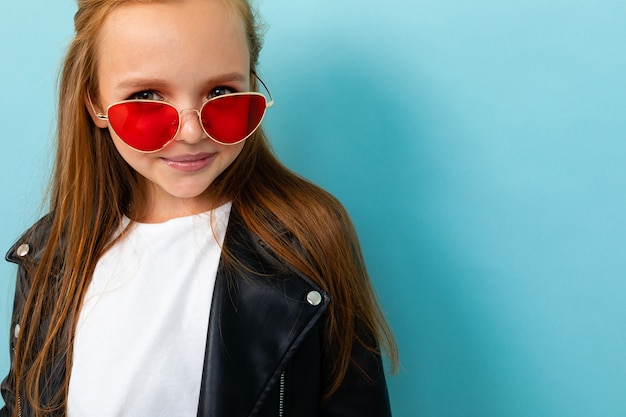 Foto retrato de medio cuerpo de una adolescente en una chaqueta negra y gafas rojas contra una pared azul