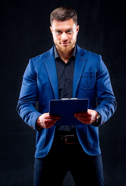 Retrato de medio cuerpo de un apuesto joven sonriente vestido con camisa oscura y traje azul con carpeta azul