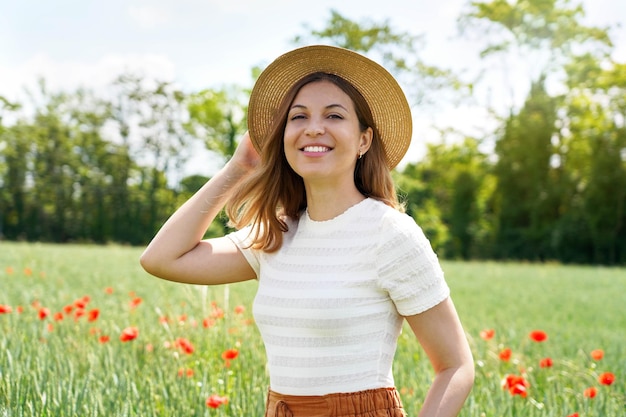 Retrato de medio cuerpo de una bella mujer joven con sombrero posando en campo de trigo con flores de amapola y mirando a cámara