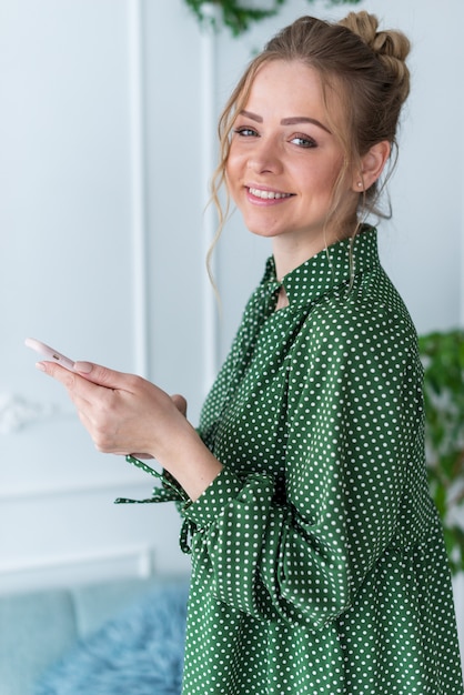 Retrato de medio cuerpo de una chica rubia de pie con un teléfono inteligente mirando a la cámara. - Imagen