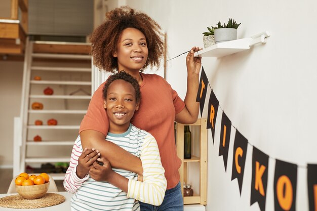 Retrato médio horizontal de uma jovem adulta feliz e seu filho alegre sorrindo para a câmera enquanto decoram a sala para o halloween