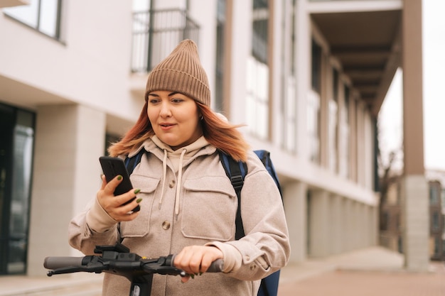 Retrato de mensajero femenino con dirección de navegación de mochila térmica en el mapa del teléfono móvil de pie en la calle de la ciudad con scooter eléctrico