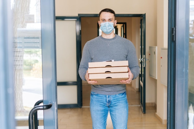 Retrato de un mensajero con máscara médica sosteniendo cajas de pizza y terminal inalámbrico POS de pago de pie en el vestíbulo de entrada del apartamento o edificio de oficinas