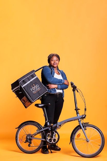 Retrato de un mensajero sonriente parado al lado de una bicicleta sosteniendo una mochila térmica de entrega para llevar, entregando el pedido del almuerzo al cliente. Trabajador de pizzería en estudio permanente sobre fondo amarillo