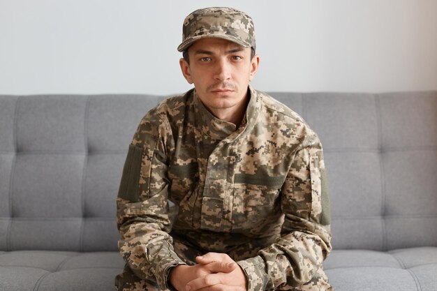 Retrato de un militar caucásico serio y tranquilo con uniforme de camuflaje y gorra sentado en un sofá y mirando a la cámara con expresión facial deprimida