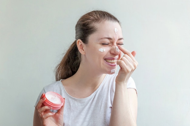 Retrato mínimo da beleza retrato da menina da jovem mulher que aplica a máscara ou a nata nutritiva branca na cara isolada na parede branca. conceito de spa cosmético orgânico de limpeza eco skincare.