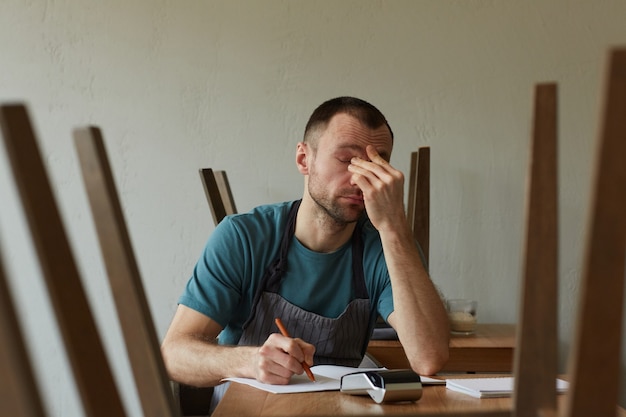 Retrato mínimo da vista frontal de um jovem exausto calculando finanças e fazendo o inventário em uma pequena loja, copie o espaço