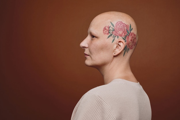 Retrato mínimo da vista lateral de uma mulher careca confiante com tatuagem na cabeça posando contra um fundo marrom no estúdio. Alopecia e conscientização do câncer, copie o espaço