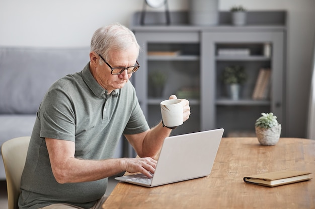 Retrato mínimo de homem sênior moderno usando o laptop em casa e tomando café, copie o espaço