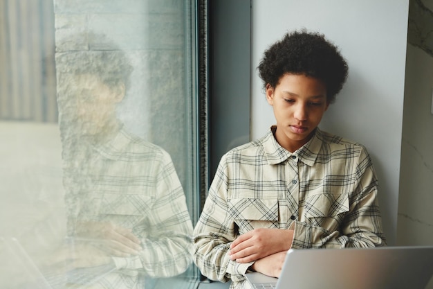 Retrato mínimo de jovem negra usando laptop enquanto está sentado pela janela no espaço de cópia da escola