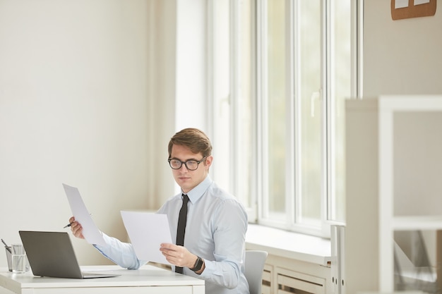 Retrato mínimo de exitoso joven empresario sosteniendo documentos mientras trabaja en el escritorio en el interior de la oficina blanca, espacio de copia