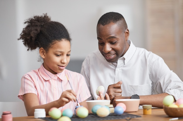 Retrato mínimo de una joven afroamericana pintando huevos de Pascua mientras disfruta de la decoración de bricolaje con el padre