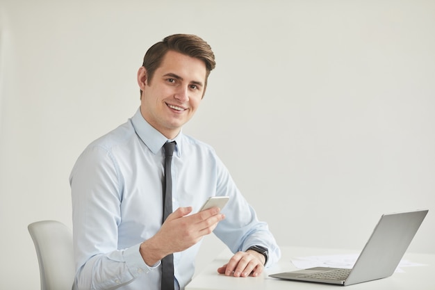 Retrato mínimo de sonriente joven empresario sosteniendo un teléfono inteligente mientras está sentado en el escritorio y mirando a la cámara trabajando en la oficina contra la pared blanca, espacio de copia