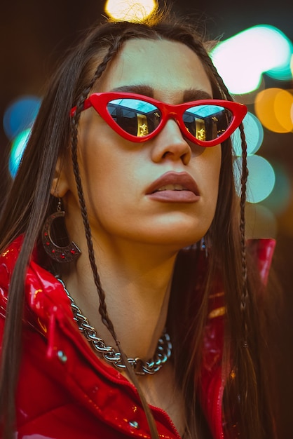 Retrato de moda al aire libre de glamour joven con cabello trenzado viste chaqueta roja y gafas de sol rojas de moda en las luces de la calle de neón