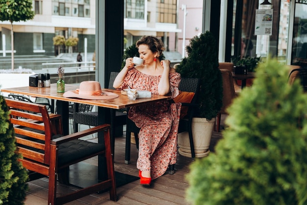 Retrato de moda al aire libre de una mujer impresionante sentada en un café Tomo café y leo un libro viejo una mujer con un vestido y un sombrero