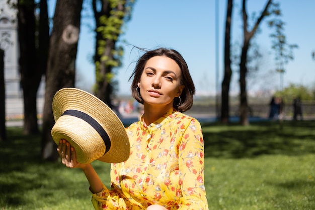 Retrato de moda al aire libre de mujer en vestido amarillo de verano y sombrero sentado en la hierba en el parque