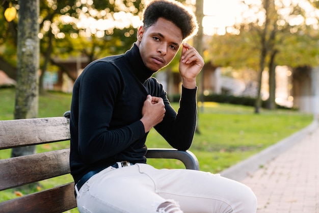 Retrato de moda de un apuesto hombre marroquí con cabello afro sentado en un banco del parque en ropa de estilo urbano en una hermosa puesta de sol