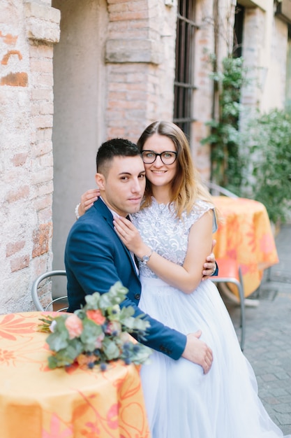 Retrato de moda de boda de la joven pareja con un bonito ramo sentado en la mesa del restaurante de la calle en Sirmione, Italia