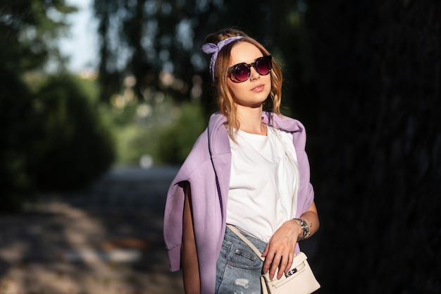 Retrato de moda brillante de sensual mujer joven hermosa con gafas de sol en una camiseta blanca y pantalones cortos con una sudadera con capucha púrpura