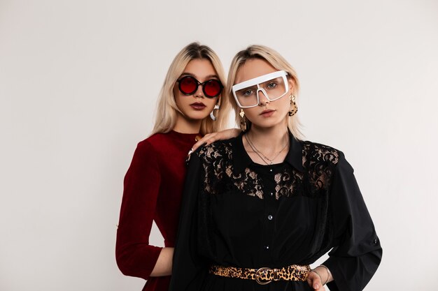 Retrato de moda dos adolescentes con gafas de colores con cabello rubio en vestidos rojo-negros de pie cerca de la pared gris vintage