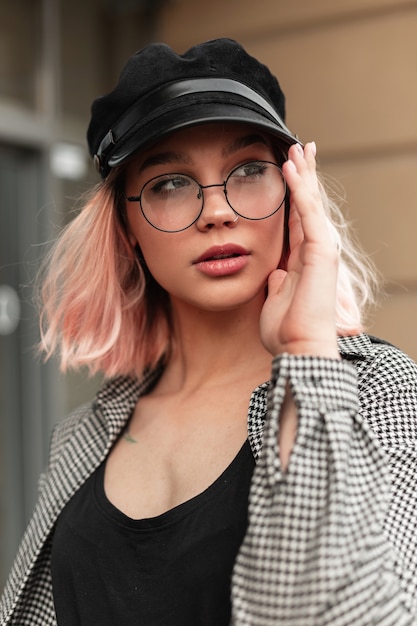 Retrato de moda elegante de una hermosa joven con cabello rosado con una camisa a cuadros y una camiseta con un sombrero se pone gafas y camina en la ciudad