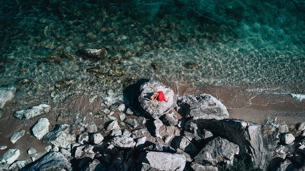 Retrato de moda de estilo de vida al aire libre de linda chica acostada en las grandes piedras en el mar Vistiendo un elegante vestido rojo largo. Solitario. Arte Fotografico. Creativo. Vista superior. Estado de ánimo romántico