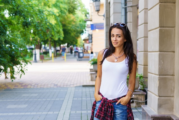 Retrato de moda de estilo de vida al aire libre de mujer joven contenta sentada al aire libre. Vistiendo jeans azules y una camiseta blanca, sonriendo, luciendo feliz y disfrutando de la vida