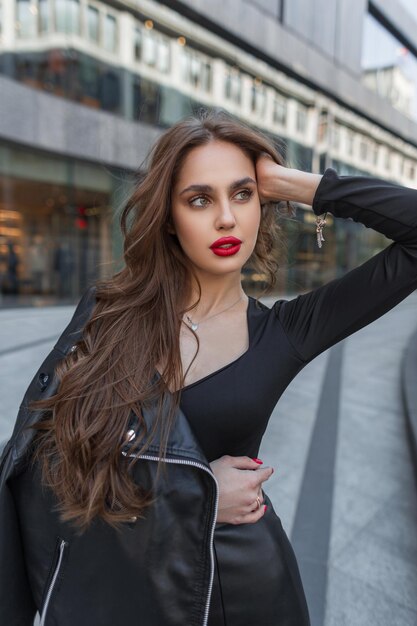 Retrato de moda femenina callejera de una hermosa dama elegante con labios rojos en ropa negra de moda con una chaqueta de cuero posando en la ciudad