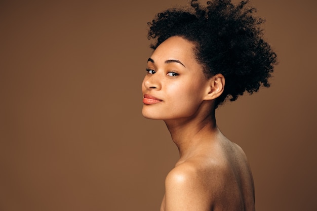 Retrato de moda de la hermosa mujer multirracial con maquillaje desnudo mirando a la cámara desde su hombro aislado sobre fondo beige