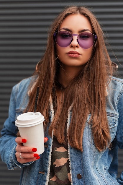 Retrato de moda Hipster de mujer joven estadounidense en elegantes gafas moradas juveniles en chaqueta de mezclilla azul de moda con una bebida caliente en las manos junto a la pared gris en la calle. Modelo de chica hermosa en la ciudad.