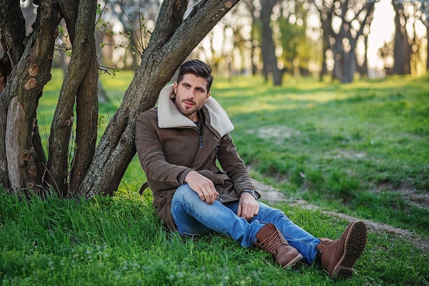 Retrato de moda joven atractivo sentado sobre la hierba verde en un parque al atardecer