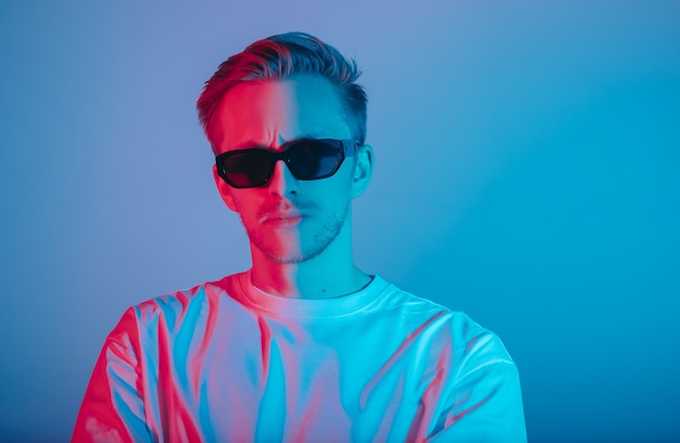 Retrato de moda joven en camiseta blanca y gafas de sol negras en luz de neón roja y azul