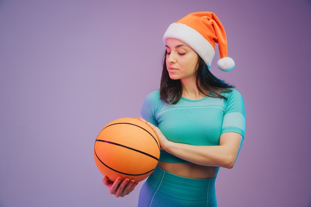 Retrato de moda de joven mujer caucásica en forma y deportiva con sombrero de Navidad con baloncesto sobre fondo degradado. Cuerpo perfecto listo para el verano. Belleza, resort, concepto de deporte.