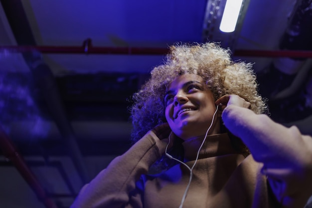 Retrato de moda joven sonriente con pelo rizado escuchando música y mirando a otro lado.