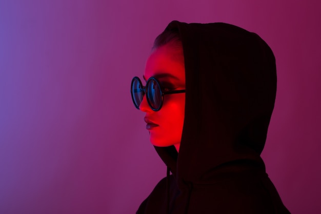 Retrato de moda joven con un suéter negro con capucha y gafas de sol redondas en luz de neón roja y azul en el estudio.